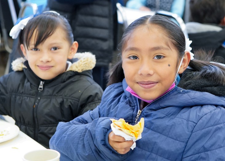 Two young girls with tan skin and brown hair smile while one holds a sandwich